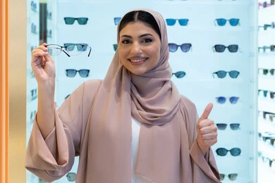 رفع اصبع الابهام للأعلى بتعابير الاعجاب، مفهوم ضعف النظر، النظر إلى الكاميرا بإيماءات السعادة، اقتناء نظارات طبية جديدة، صورة مقربة لشابة عربية خليجية سعودية ترتدي عباءة تقوم باختيار نظارة ملائمة لملامح وجهها، امرأة إماراتية تقوم بمعاينة النظارات