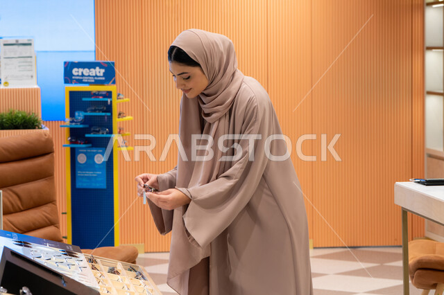 مفهوم الاهتمام بالمظهر الخارجي، مواسم التخفيضات في الإمارات، مواكبة آخر صيحات الموضة والجمال، شابة عربية خليجية اماراتية محجبة ترتدي عباءة تقوم باختيار نظارة شمسية ملائمة لملامح الوجه، امرأة سعودية تقوم بمعاينة النظارات