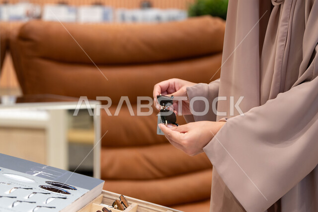 الاستفادة من العروض والتخفيضات، الاهتمام بالمظهر الخارجي، مواكبة آخر صيحات الموضة والجمال، صورة مقربة لأيدي شابة عربية خليجية اماراتية  تقوم باختيار نظارة شمسية ملائمة لملامح الوجه، امرأة سعودية تقوم بمعاينة النظارات