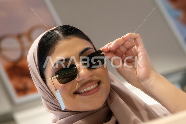 تجربة النظارات الشمسيه قبل الشراء، التسوق في المولات والمتاجر الإماراتية، انواع وماركات عالميه، سيدة سعودية تقوم باختيار نظارة شمسيه مناسبة، صورة مقربة لامرأة عربية خليجية إماراتية محجبة مبتسمة ترتدي عباءة تقوم بقياس نظاره بإيماءات السعادة والسرور