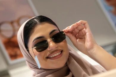 تجربة النظارات الشمسيه قبل الشراء، التسوق في المولات والمتاجر الإماراتية، انواع وماركات عالميه، سيدة سعودية تقوم باختيار نظارة شمسيه مناسبة، صورة مقربة لامرأة عربية خليجية إماراتية محجبة مبتسمة ترتدي عباءة تقوم بقياس نظاره بإيماءات السعادة والسرور