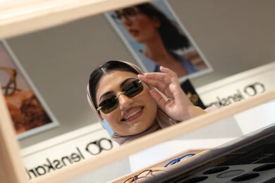 التسوق في المولات والمتاجر الإماراتية، انواع وماركات عالميه، ارتداء النظارات الشمسيه قبل الشراء، سيدة سعودية تقوم باختيار نظارة شمسيه مناسبة، صورة مقربة لامرأة عربية خليجية إماراتية محجبة مبتسمة ترتدي عباءة تقوم بقياس نظاره بإيماءات السعادة والسرور