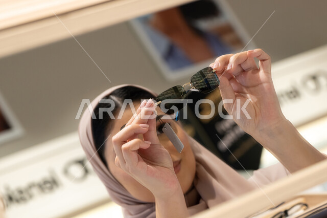 Trying on sunglasses before buying, shopping in malls and stores in the UAE, international types and brands, a Saudi woman choosing suitable sunglasses, a close-up photo of a smiling veiled Emirati Gulf Arab woman wearing an abaya trying on her glasses with gestures of happiness and pleasure