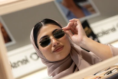 متعة التسوق والشراء، سيدة سعودية تقوم باختيار نظارة شمسيه مناسبة، ارتداء النظارات الشمسيه قبل الشراء، صورة مقربة لامرأة عربية خليجية إماراتية محجبة ترتدي عباءة تقوم بقياس نظاره بإيماءات الثقة بالنفس، ارفف عرض المنتجات داخل المحل، انواع وماركات عالميه