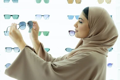 Keeping up with the latest fashion and beauty trends, the latest stylish women's accessories, the joy of shopping in the UAE malls, a Saudi woman examining colored glasses, a side view of a veiled young Emirati Gulf Arab woman wearing an abaya choosing sunglasses that suit her facial features