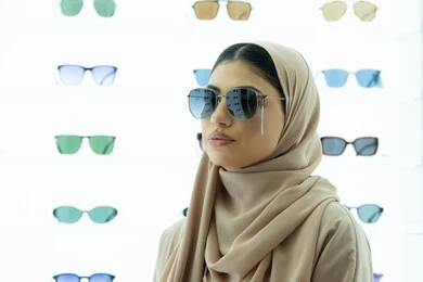 متعة التسوق والشراء، سيدة سعودية تقوم باختيار نظارة شمسيه مناسبة، ارتداء النظارات الشمسيه قبل الشراء، صورة مقربة لامرأة عربية خليجية إماراتية محجبة ترتدي عباءة تقوم بقياس نظاره بإيماءات الثقة بالنفس، ارفف عرض المنتجات داخل المحل، انواع وماركات عالميه