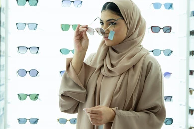 الاهتمام بالمظهر الخارجي، اكسسوارات نسائية عصرية، التأثير الإيجابي للتسوق على المرأة، تجربة النظارات الشمسيه قبل الشراء، أرفف عرض المنتجات في المتجر، شابة اماراتية تقوم باختيار نظارة شمسيه مناسبة، سيدة عربية خليجية سعودية محجبة ترتدي عباءة تقوم بقياس نظاره