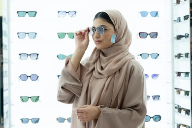 اكسسوارات نسائية عصرية، التأثير الإيجابي للتسوق على المرأة، تجربة النظارات الشمسيه قبل الشراء، أرفف عرض المنتجات في المتجر، شابة اماراتية تقوم باختيار نظارة شمسيه مناسبة، سيدة عربية خليجية سعودية محجبة ترتدي عباءة تقوم بقياس نظاره