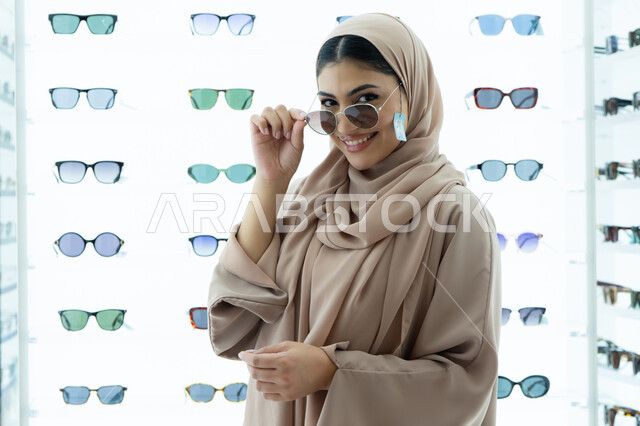 النظر إلى الكاميرا بتعابير السرور، تجربة النظارات الشمسيه قبل الشراء، أرفف عرض المنتجات في المتجر، متعة التسوق في المولات الإماراتية، سيدة سعودية تقوم باختيار نظارة شمسيه مناسبة، صورة مقربة لامرأة عربية خليجية إماراتية محجبة مبتسمة ترتدي عباءة تقوم بقياس نظاره