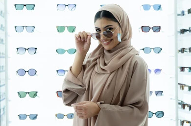 النظر إلى الكاميرا بتعابير السرور، تجربة النظارات الشمسيه قبل الشراء، أرفف عرض المنتجات في المتجر، متعة التسوق في المولات الإماراتية، سيدة سعودية تقوم باختيار نظارة شمسيه مناسبة، صورة مقربة لامرأة عربية خليجية إماراتية محجبة مبتسمة ترتدي عباءة تقوم بقياس نظاره