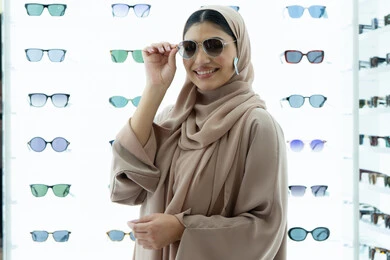 التأثير الإيجابي للتسوق على المرأة، النظر إلى الكاميرا بتعابير السرور، تجربة النظارات الشمسيه قبل الشراء، أرفف عرض المنتجات في المتجر، شابة سعودية تقوم باختيار نظارة شمسيه مناسبة، صورة مقربة لسيدة عربية خليجية إماراتية محجبة مبتسمة ترتدي عباءة تقوم بقياس نظاره