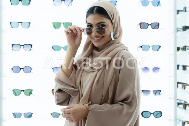 متعة التسوق في المولات الإماراتية، النظر إلى الكاميرا بتعابير السرور، تجربة النظارات الشمسيه قبل الشراء، أرفف عرض المنتجات في المتجر، شابة سعودية تقوم باختيار نظارة شمسيه مناسبة، صورة مقربة لامرأة عربية خليجية إماراتية محجبة مبتسمة ترتدي عباءة تقوم بقياس نظاره