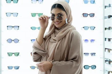 متعة التسوق في المولات الإماراتية، النظر إلى الكاميرا بتعابير السرور، تجربة النظارات الشمسيه قبل الشراء، أرفف عرض المنتجات في المتجر، شابة سعودية تقوم باختيار نظارة شمسيه مناسبة، صورة مقربة لامرأة عربية خليجية إماراتية محجبة مبتسمة ترتدي عباءة تقوم بقياس نظاره