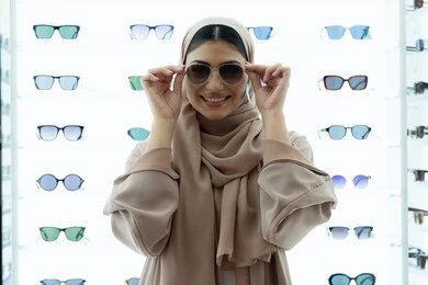 أرفف عرض المنتجات في المتجر، التسوق في المولات الإماراتية، تجربة النظارات الشمسيه قبل الشراء، انواع وماركات عالميه، سيدة سعودية تقوم باختيار نظارة شمسيه مناسبة، صورة مقربة لامرأة عربية خليجية إماراتية محجبة مبتسمة ترتدي عباءة تقوم بقياس نظاره بإيماءات السعادة