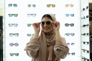 عرض المنتجات على الأرفف، التسوق في المولات الإماراتية، تجربة النظارات الشمسيه قبل الشراء، انواع وماركات عالميه، سيدة سعودية تقوم باختيار نظارة شمسيه مناسبة، صورة مقربة لامرأة عربية خليجية إماراتية محجبة مبتسمة ترتدي عباءة تقوم بقياس نظاره بإيماءات السعادة