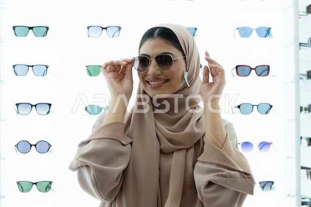 Trying on sunglasses before buying, display shelves in a store, shopping in UAE malls, international brands and types, a Saudi woman choosing suitable sunglasses, a close-up of a smiling veiled Emirati Gulf Arab woman wearing an abaya trying on glasses with happy gestures
