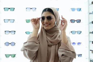تجربة النظارات الشمسيه قبل الشراء، أرفف عرض المنتجات في المتجر، التسوق في المولات الإماراتية، انواع وماركات عالميه، سيدة سعودية تقوم باختيار نظارة شمسيه مناسبة، صورة مقربة لامرأة عربية خليجية إماراتية محجبة مبتسمة ترتدي عباءة تقوم بقياس نظاره بإيماءات السعادة