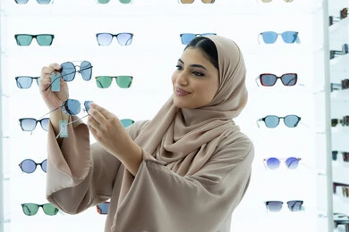 شراء اكسسوارات نسائية عصرية أنيقة، متابعة آخر صيحات الموضة والجمال، متعة التسوق في مولات الإمارات، امرأة سعودية تقوم بمعاينة النظارات الملونة، شابة عربية خليجية اماراتية محجبة ترتدي عباءة تقوم باختيار نظارة شمسية ملائمة لملامح الوجه