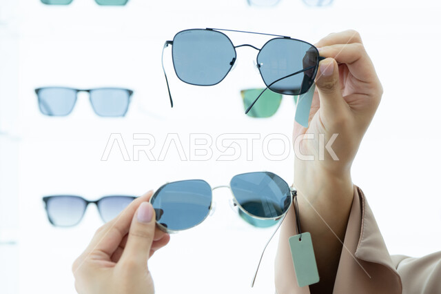 Sunglasses exhibition, taking advantage of offers and discounts, keeping up with the latest fashion and beauty trends, a close-up of the hands of a young Arab Gulf Emirati woman choosing sunglasses that suit her facial features, a Saudi woman examining glasses