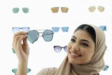 محل لبيع أحدث الاكسسوارات النسائية، المتعة بالتسوق في مولات الإمارات، مواكبة آخر صيحات الموضة والجمال، صورة مقربة لشابة عربية خليجية اماراتية محجبة ترتدي عباءة تقوم باختيار نظارة شمسية ملائمة لملامح الوجه،  امرأة سعودية تقوم بمعاينة النظارات بتعابير السعادة