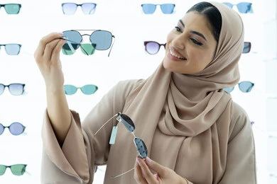 السعادة والمتعة بالتسوق في مولات الإمارات، مواكبة آخر صيحات الموضة والجمال، أحدث الاكسسوارات النسائية العصرية الأنيقة، صورة مقربة لشابة عربية خليجية اماراتية محجبة ترتدي عباءة تقوم باختيار نظارة شمسية ملائمة لملامح الوجه،  امرأة سعودية تقوم بمعاينة النظارات