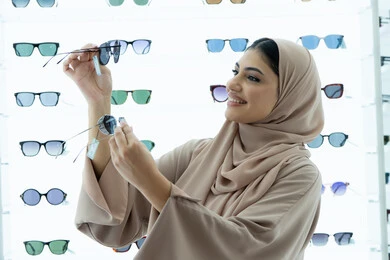 السرور والمتعة بالتسوق في مولات الإمارات، مواكبة آخر صيحات الموضة والجمال، أحدث الاكسسوارات النسائية العصرية الأنيقة، صورة مقربة لشابة عربية خليجية اماراتية محجبة ترتدي عباءة تقوم باختيار نظارة شمسية ملائمة لملامح الوجه،  امرأة سعودية تقوم بمعاينة النظارات