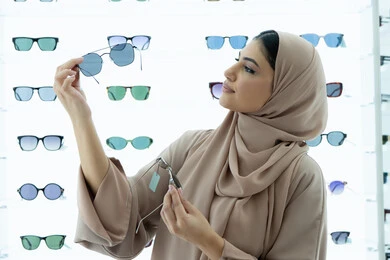 متابعة آخر صيحات الموضة والجمال، متعة التسوق في مولات الإمارات، اكسسوارات نسائية عصرية أنيقة، صورة مقربة لامرأة سعودية تقوم بمعاينة النظارات الملونة، شابة عربية خليجية اماراتية محجبة ترتدي عباءة تقوم باختيار نظارة شمسية ملائمة لملامح الوجه