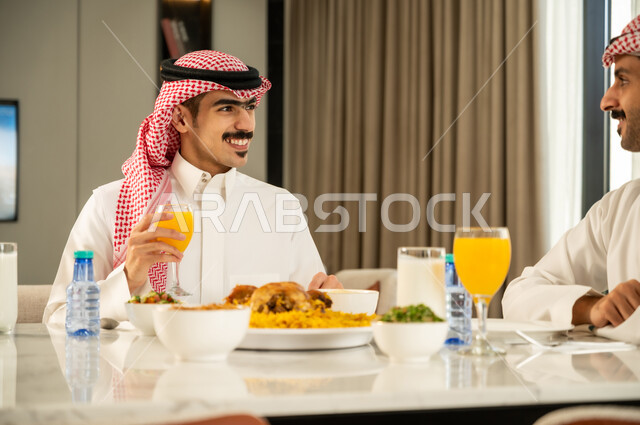 تقديم الكبسه السعوديه على المائده، إيماءات السعادة و السرور بقضاء وقت ممتع مع الأصدقاء، دعوة العائلة لتناول وجبة شهيه, حسن إكرام واستقبال الضيف، الاجتماع على طاولة الطعام، صورة مقربة لصديقين عربيين خليجيين سعوديين يرتديان الزي التقليدي يتناولان الغداء