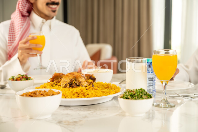 تقديم الكبسه السعوديه على المائده، إيماءات السعادة و السرور بقضاء وقت ممتع مع الأصدقاء، دعوة العائلة لتناول وجبة شهيه, حسن إكرام واستقبال الضيف، الاجتماع على طاولة الطعام، صورة مقربة لصديقين عربيين خليجيين سعوديين يرتديان الزي التقليدي يتناولان الغداء