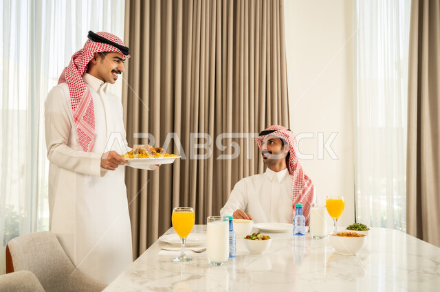 Gathering at the dining table, inviting the family to a delicious meal, a Saudi Arabian Gulf man wearing the traditional thobe and shemagh serving Saudi Kabsa to his friend at the table, gestures of happiness and pleasure in spending a pleasant time with friends, good hospitality and reception of the guest, a popular Saudi Gulf dish