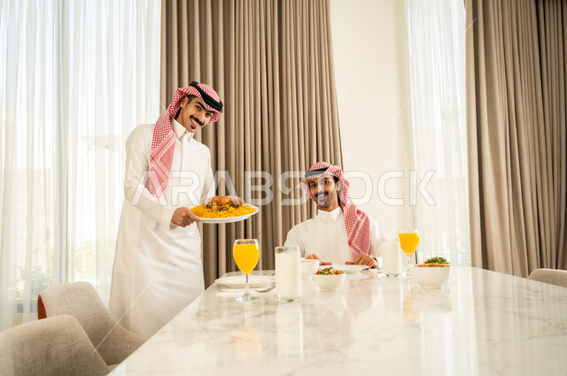 النظر إلى الكاميرا بإيماءات السعادة و السرور، الاجتماع على طاولة الطعام، دعوة العائلة لتناول وجبة شهيه, رجل عربي خليجي سعودي يرتدي الثوب التقليدي والشماغ يقوم بتقديم الكبسه السعوديه لصديقه على المائده، قضاء وقت ممتع مع الأصدقاء, حسن إكرام واستقبال الضيف