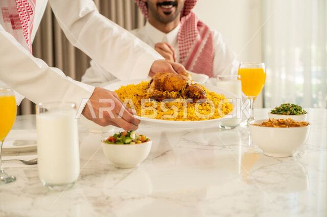الاجتماع على طاولة الطعام، دعوة العائلة لتناول وجبة شهيه, صورة مقربة لرجل عربي خليجي سعودي يرتدي الثوب التقليدي والشماغ يقوم بتقديم الكبسه السعوديه لصديقه على المائده، قضاء وقت ممتع مع الأصدقاء, حسن إكرام واستقبال الضيف