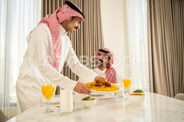 الاجتماع على طاولة الطعام، دعوة العائلة لتناول وجبة شهيه, رجل عربي خليجي سعودي يرتدي الثوب التقليدي والشماغ يقوم بتقديم الكبسه السعوديه لصديقه على المائده، إيماءات السعادة و السرور بقضاء وقت ممتع مع الأصدقاء, حسن إكرام واستقبال الضيف، أكلة شعبية خليجية سعودية