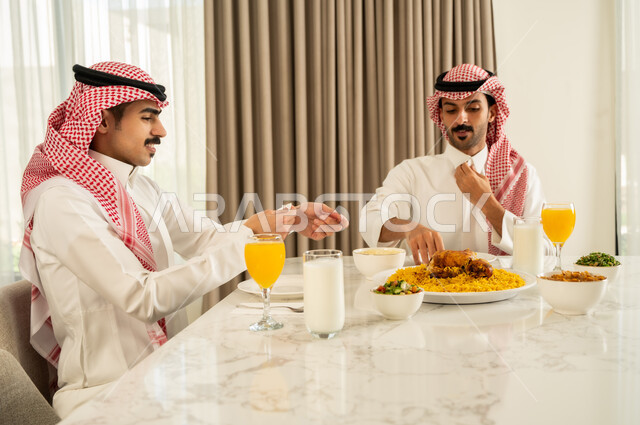 الاجتماع على طاولة الطعام ، دعوة العائلة لتناول وجبة شهيه، تقديم الكبسه السعوديه على المائده, صديقان عربيان خليجيان سعوديان يرتديان الثوب التقليدي و الشماغ يقومان بتناول المأكولات بإيماءات السعادة و السرور, حسن إكرام واستقبال الضيف