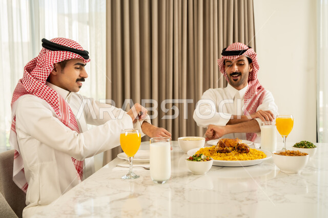 الاجتماع على طاولة الطعام ، دعوة العائلة لتناول وجبة شهيه، تقديم الكبسه السعوديه على المائده, صديقان عربيان خليجيان سعوديان يرتديان الثوب التقليدي و الشماغ يقومان بتناول المأكولات بإيماءات السعادة و السرور, حسن إكرام واستقبال الضيف