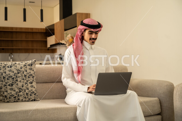 Watching movies and series, browsing the internet and working remotely, working hard and completing the required tasks, using modern technology, mastering computer and information technology jobs, a Saudi Arabian Gulf man wearing traditional clothing sitting on the sofa in the living room and using the laptop
