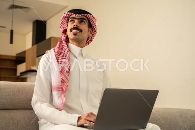 احتراف وظائف الحاسوب وتكنولوجيا المعلومات، النظر إلى شيء ما، تصفح مواقع الانترنت والعمل عن بعد، الاجتهاد وانجاز الاعمال المطلوبة، الاعجاب بالتقنية الحديثة، رجل عربي خليجي سعودي يرتدي الزي التقليدي يجلس على الأريكة في غرفة المعيشة ويقوم باستخدام اللابتوب