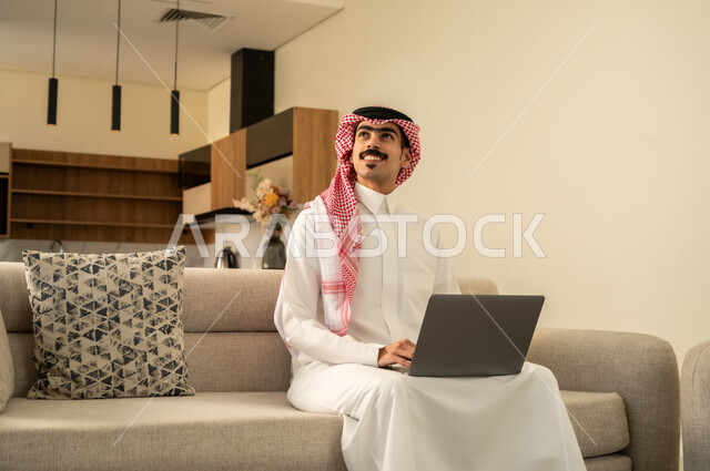 Looking at something, browsing the internet and working remotely, working hard and completing the required tasks, admiring modern technology, professionalism in computer and information technology jobs, a Saudi Arabian Gulf man wearing traditional clothing sitting on the sofa in the living room and using the laptop