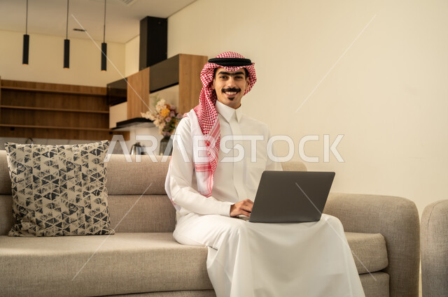 احتراف وظائف الحاسوب وتكنولوجيا المعلومات، النظر إلى الكاميرا بتعابير السرور، تصفح مواقع الانترنت والعمل عن بعد، الاجتهاد وانجاز الاعمال المطلوبة، الاعجاب بالتقنية الحديثة، رجل عربي خليجي سعودي يرتدي الزي التقليدي يجلس على الأريكة في غرفة المعيشة ويستخدم اللابتوب