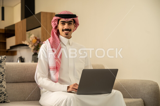 الاعجاب بالتقنية الحديثة، احتراف وظائف الحاسوب وتكنولوجيا المعلومات، النظر إلى الكاميرا بتعابير السرور، تصفح مواقع الانترنت والعمل عن بعد، الاجتهاد وانجاز الاعمال المطلوبة، رجل عربي خليجي سعودي يرتدي الزي التقليدي يجلس على الأريكة في غرفة المعيشة ويستخدم اللابتوب
