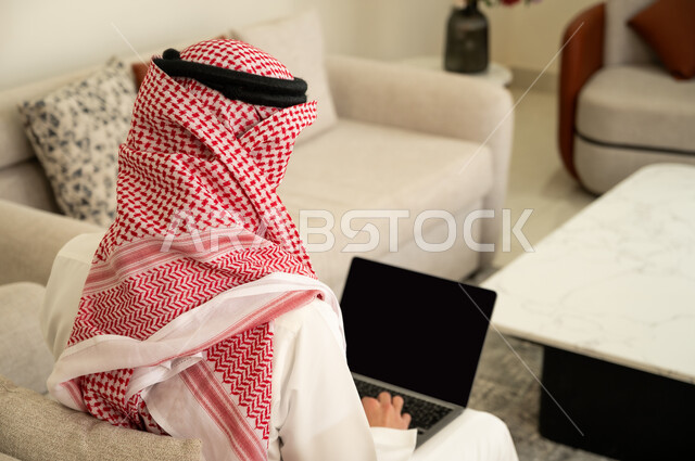 Hard work and completion of required tasks, admiration of modern technology, professionalism in computer and information technology jobs, surfing the internet and working remotely, close-up photo from the back of a Saudi Arabian Gulf man wearing traditional clothing sitting on the sofa in the living room and using a laptop