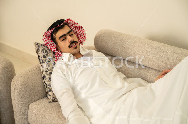 النوم بعمق من الارهاق والتعب، رجل عربي خليجي سعودي يرتدي الثوب والشماغ يقوم بالاستلقاء على الاريكة في منزله، اخذ قيلولة و قسط من الراحة، الاحساس بالكسل والخمول والحاجة إلى النوم، الاسترخاء والاستمتاع بالأجواء الهادئة