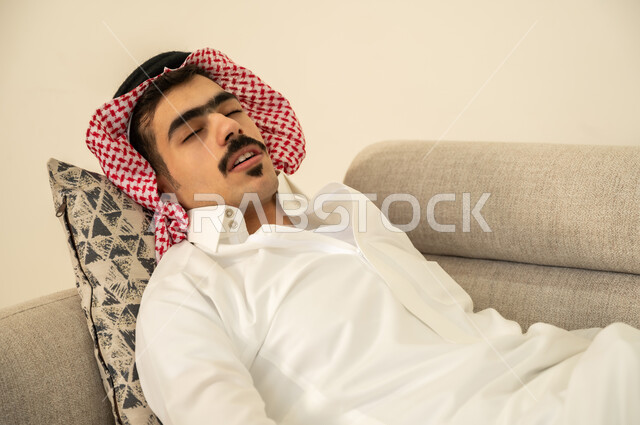 اخذ قيلولة و قسط من الراحة، الاحساس بالكسل والخمول والحاجة إلى الاسترخاء، النوم بعمق من الارهاق والتعب، صورة مقربة لرجل عربي خليجي سعودي يرتدي الثوب والشماغ يقوم بالاستلقاء على الاريكة في منزله، الاستمتاع بالأجواء الهادئة