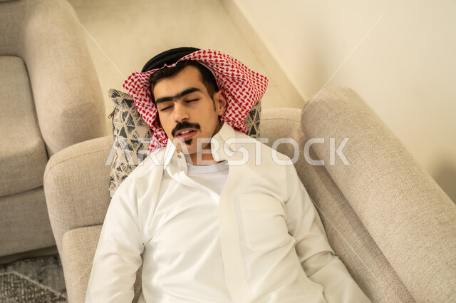 اخذ قيلولة و قسط من الراحة، الاحساس بالكسل والخمول والحاجة إلى الاسترخاء، النوم بعمق من الارهاق والتعب، صورة مقربة لرجل عربي خليجي سعودي يرتدي الثوب والشماغ يقوم بالاستلقاء على الاريكة في منزله، الاستمتاع بالأجواء الهادئة