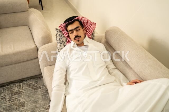 النوم بعمق من الارهاق والتعب، رجل عربي خليجي سعودي يرتدي الثوب والشماغ يقوم بالاستلقاء على الاريكة في منزله، اخذ قيلولة و قسط من الراحة، الاحساس بالكسل والخمول والحاجة إلى النوم، الاسترخاء والاستمتاع بالأجواء الهادئة