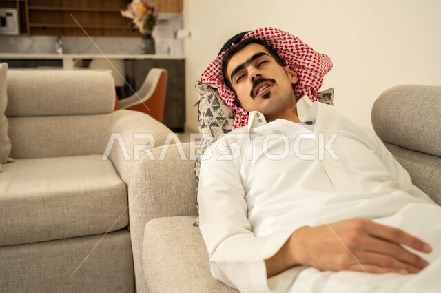 الاحساس بالكسل والخمول والحاجة إلى الاسترخاء، النوم بعمق من الارهاق والتعب، رجل عربي خليجي سعودي يرتدي الثوب والشماغ يقوم بالاستلقاء على الاريكة في منزله، اخذ قيلولة و قسط من الراحة، الاستمتاع بالأجواء الهادئة