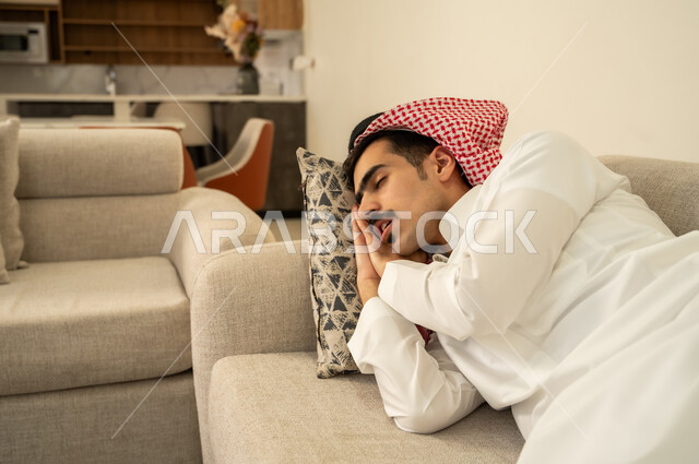Sleeping deeply from exhaustion and fatigue, a Saudi Arabian Gulf man wearing a thobe and shemagh lying on the sofa in his home, taking a nap and resting, feeling lazy and sluggish and in need of sleep, relaxing and enjoying the calm atmosphere