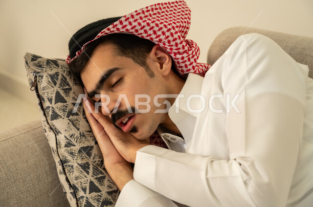 اخذ قيلولة و قسط من الراحة، الاحساس بالكسل والخمول والحاجة إلى الاسترخاء، النوم بعمق من الارهاق والتعب، صورة مقربة لرجل عربي خليجي سعودي يرتدي الثوب والشماغ يقوم بالاستلقاء على الاريكة في منزله، الاستمتاع بالأجواء الهادئة