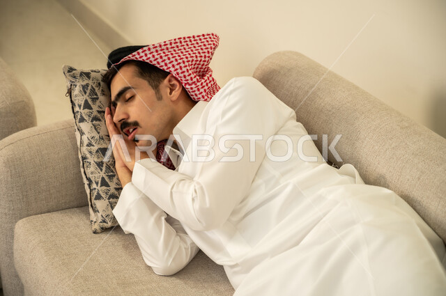الاحساس بالكسل والخمول والحاجة إلى الاسترخاء، النوم بعمق من الارهاق والتعب، رجل عربي خليجي سعودي يرتدي الثوب والشماغ يقوم بالاستلقاء على الاريكة في منزله، اخذ قيلولة و قسط من الراحة، الاستمتاع بالأجواء الهادئة