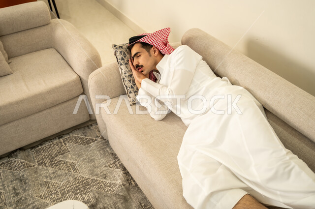 النوم بعمق من الارهاق والتعب، رجل عربي خليجي سعودي يرتدي الثوب والشماغ يقوم بالاستلقاء على الاريكة في منزله، اخذ قيلولة و قسط من الراحة، الاحساس بالكسل والخمول والحاجة إلى النوم، الاسترخاء والاستمتاع بالأجواء الهادئة